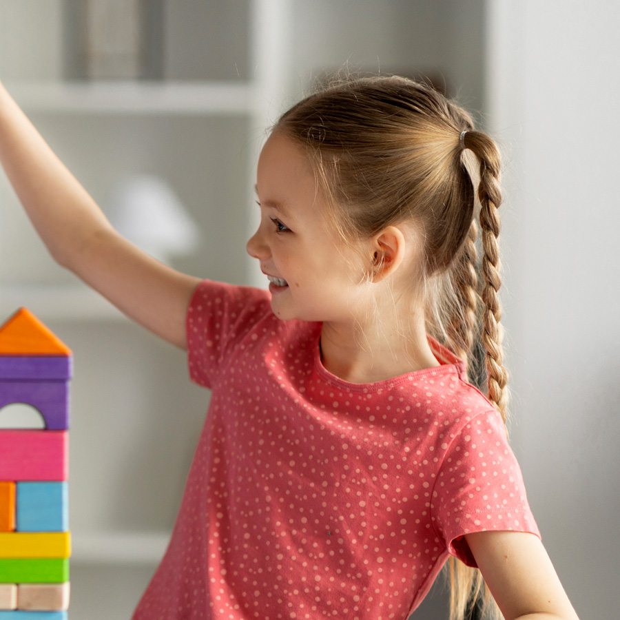 A young girl smiles while playing with colorful building blocks indoors.