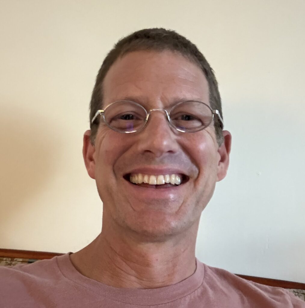 A smiling man wearing glasses and a casual shirt.