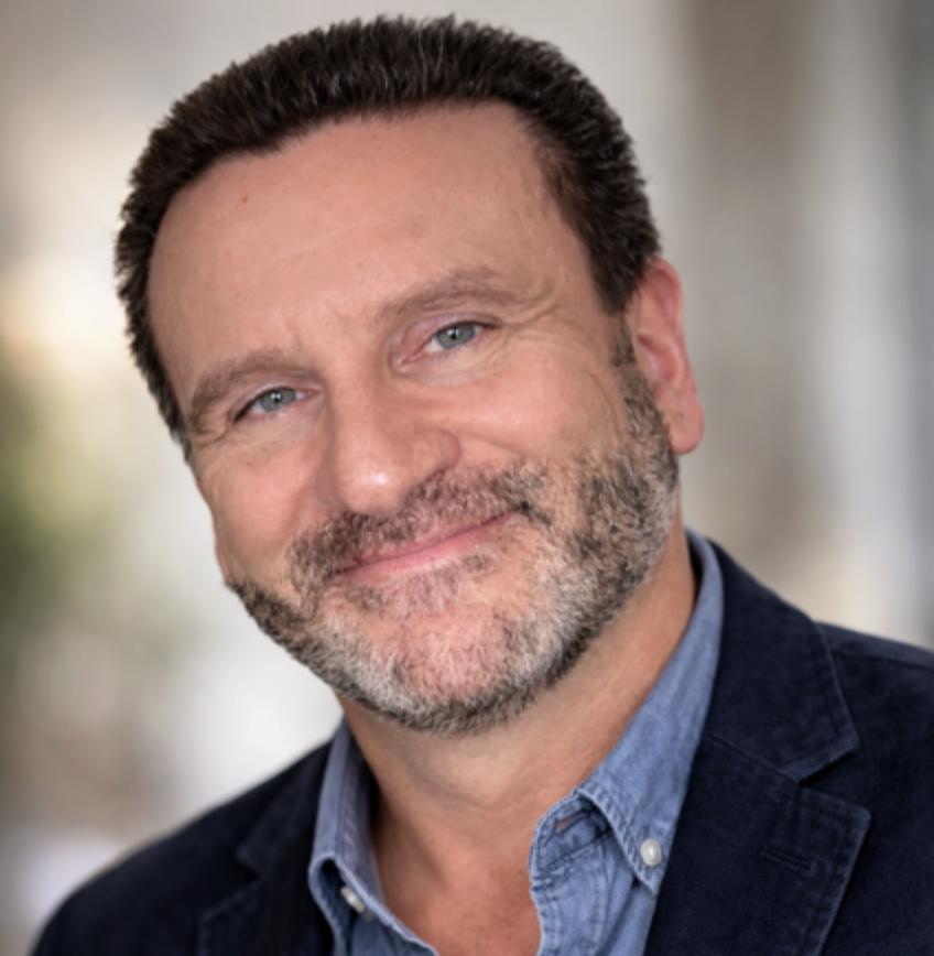 Smiling middle-aged man with curly hair and beard in a blue shirt and dark jacket.